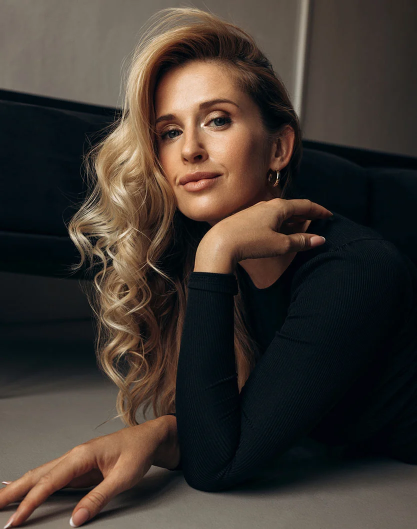 A stunning portrait of a blonde woman with wavy hair, wearing a black long-sleeved shirt. She is lying on the floor, propping her head up with one hand while looking confidently at the camera. - Eyelid Surgery in Los Angeles, CA
