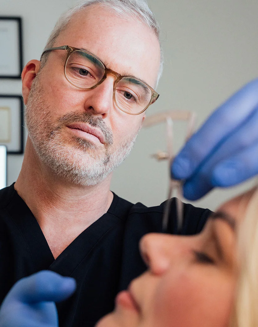 A male plastic surgeon, Dr. Mather, wearing blue surgical gloves, glasses, and black scrubs, examines a client's nose from above, holding a measuring tool. The client's face is partially visible in the foreground. - Rhinoplasty in Los Angeles, CA